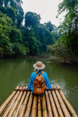 Tayland 'da Khao Sok' ta bir gezgin bir bambu salında dinleniyor. Sahne, sakin suları kaplayan yoğun ve canlı ormanlarla huzurlu atmosferi yakalıyor, macera ve yansımayı davet ediyor..