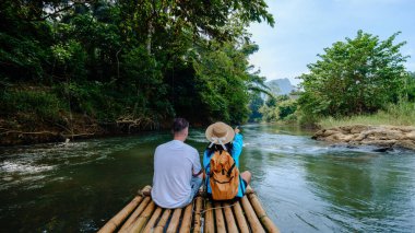 İki gezgin Tayland 'daki Khao Sok Ulusal Parkı' nda yemyeşil yemyeşil bir nehirde huzurlu bir bambu rafting deneyiminin tadını çıkarıyor. Atmosfer sakin ve resim gibi..