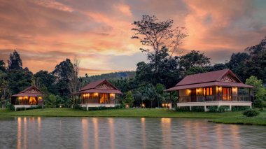 Bungalovlar Tayland, Khao Sok 'ta sakin bir gölün yanında durarak huzur içinde geri çekiliyorlar. Canlı günbatımı gökyüzü sıcaklık katar, doğaya mükemmel bir kaçış sunar güzellik.