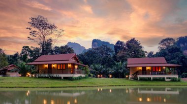 Büyüleyici ahşap bungalovlar, Tayland, Khao Sok 'ta canlı bir günbatımı sırasında yemyeşil ve dağlarla çevrili bir gölün huzurlu sularına yansıyor. Doğanın güzelliği parıldıyor.