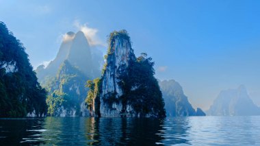 Kireçtaşı zirveleri, bereketli yeşilliklerle çevrili Cheow Lan Gölü 'nün sakin suları üzerinde görkemli bir şekilde yükselir. Bu sakin manzara, Tayland 'daki Khao Sok Ulusal Parkı' nın doğal güzelliğini gözler önüne seriyor..