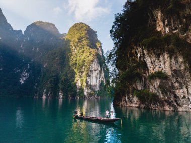 Khao Sok, Tayland 'daki Cheow Lan Gölü' nde gün doğumunda uzun kuyruklu bir teknenin önünde bir çift. Zümrüt suları, maceralar için mükemmel bir zemin sunan yüksek kireçtaşı kayalıklarıyla çevrilidir. 