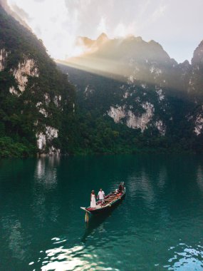 İki yolcu, Cheow Lan Gölü 'nün çarpıcı manzarasının ortasında geleneksel bir teknede huzurlu bir gezintinin tadını çıkarıyor. Khao Sok, Tayland, güneş doğarken uzun kuyruklu bir teknenin önünde bir çift.