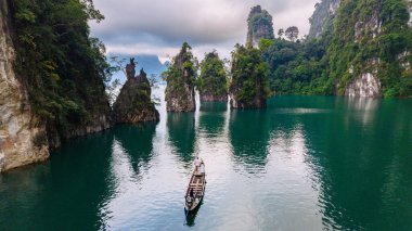 Geleneksel bir ahşap tekne Khao Sok Tayland 'ın zümrüt sularında süzülürken Cheow Lan Gölü' nde sakin bir manzara gözler önüne seriliyor.