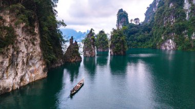 Zümrüt sularının yükselen kireçtaşı kayalıkları yansıttığı Cheow Lan Gölü 'nün çarpıcı güzelliğini keşfedin. Geleneksel bir tekne huzur içinde huzurlu bir şekilde süzülüyor, Khao Sok Tayland