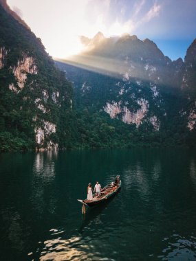 Khao Sok, Tayland 'daki Khao Lan Gölü' nün dingin güzelliğini tecrübe edin. Yüksek kireçtaşı kayalıklarının yemyeşil ormanlarla buluştuğu yer. Gün doğumunda uzun kuyruklu bir teknenin önünde bir çift.