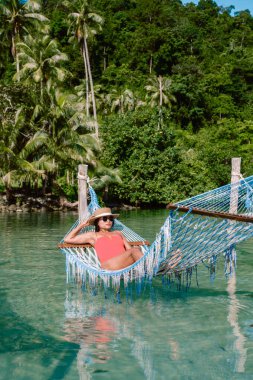Tayland 'da Koh Kood Adası' nda yemyeşil sularla çevrili sakin suların üzerindeki renkli bir hamakta dinlenen bir kadın. Bu sahne, Tayland 'daki hayat ve sükunetin özünü yakalar.