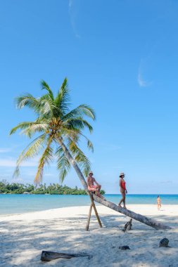 Çeşitli bir çift, Tayland 'daki Koh Kood Adası' nda eğimli bir palmiye ağacında güneşli bir günün tadını çıkarıyor. Berrak mavi sular ve yumuşak kum rahatlamak için mükemmel bir tropikal cennet atmosferi yaratır..