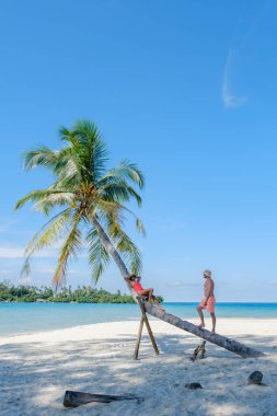 Çeşitli Kafkas erkekleri ve Asyalı kadınlar Koh Kood Adası 'ndaki kumsalda günün tadını çıkarıyorlar. Biri eğilmiş bir palmiye ağacına tırmanırken diğeri Tayland 'da okyanusla çevrili kıyıda dinleniyor.