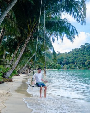 Bir adam Koh Kood Adası 'nın kıyısında salıncakta huzurlu bir anın tadını çıkarıyor. Kumlu sahil ve turkuaz suları çevreleyen yemyeşil palmiye ağaçları, sakin bir tropikal atmosfer yaratıyor..