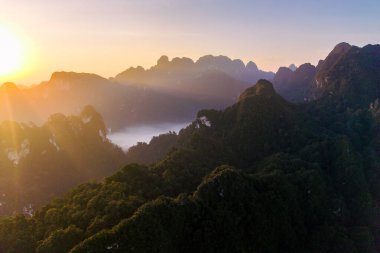 Şafak vakti Khao Sok, Tayland 'ın dingin güzelliğini deneyimleyin. Canlı renklerin gökyüzünü boyadığı ve sislerin yemyeşil dağları sardığı, ruhları büyüleyen büyülü bir manzara yaratan yer..