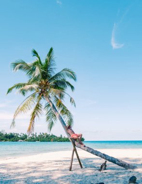 Tayland 'daki Koh Kood Adası' nda bir adamın eğimli bir palmiye ağacına uzandığı sakin bir sahne sergileniyor. Parlak mavi gökyüzü ve turkuaz sular mükemmel bir tropikal kaçış yaratıyor, rahatlamaları davet ediyor..