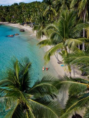 Tayland 'daki Koh Kood Adası' nın dingin güzelliğini keşfedin. Titrek palmiyeler rüzgarda hafifçe sallanıyor. İki kişi kumsalda geziniyor, sakin turkuaz suların tadını çıkarıyorlar..