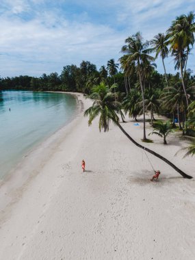 Koh Kood Adası 'nda yumuşak beyaz kum ve gür palmiye ağaçlarıyla sakin bir plaj manzarası. Tayland 'da bir plajda, palmiye ağacının altında salıncakta sallanan bir çift adam ve bir kadın.