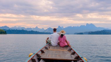 Bir çift gün batımında büyüleyici kireçtaşı dağlarıyla çevrili Cheow Lan Gölü 'nde sakin bir tekne yolculuğunun tadını çıkarıyor. Khao Sok, Tayland