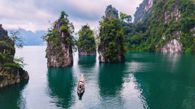 Cheow Lan Gölü 'nün sakin manzarası, yemyeşil ağaçlarla çevrili yüksek kireçtaşı kayalıkları sergiliyor. Geleneksel bir tekne zümrüt sularında usulca süzülür, doğanın güzelliğini gösterir..