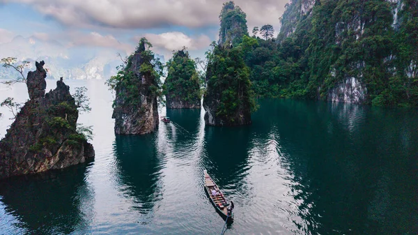Khao Sok, Tayland 'daki Cheow Lan Gölü' nün sakin sularını keşfedin. Yükselen kireçtaşı kayalıkları bulutlu bir gökyüzünün altındaki sakin zümrüt sularda süzülen uzun kuyruklu botlar için görkemli bir zemin sağlar..