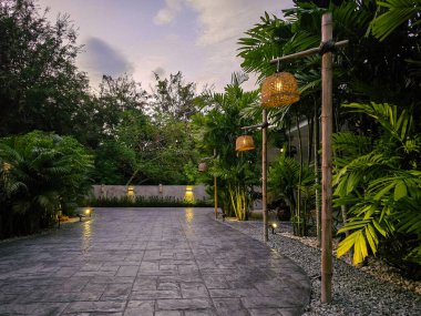 Bangkok 'taki sakin bir akşam sahnesinde, tropik yeşilliklerle çevrili çarpıcı bir beton yol bulunuyor. Yumuşak ışıklar, güneş bu huzurlu inzivada batarken canlı bitkileri aydınlatıyor..