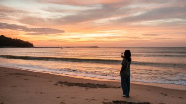 Tayland 'ın Baan Krood Sahili' nde sakin bir günbatımı, bir insan kıyıda durup anın güzelliğini izlerken gökyüzünde canlı renkler sergiliyor. Dalgalar yavaşça kumsala vurur..
