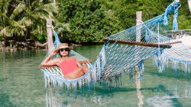 Koh Kood Adası 'nın sakin suları üzerinde asılı renkli bir hamakta uzanan bir kadın. Bereketli yeşilliklerle çevrili bu tropik cennetteki dingin güzelliğin ve ılık güneşin tadını çıkarıyor..