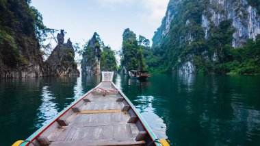 Tahta bir tekne, dramatik kireçtaşı tepeleri ve yemyeşil yeşilliklerle çevrili Cheow Lan Gölü 'nün sakin sularında süzülüyor. Khao Sok, Tayland