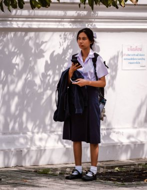 Bangkok, Tayland, 19 Kasım 2025, genç bir öğrenci duvarın yanında duruyor, ceketini tutarken telefonunu kontrol ediyor. Sıcak güneş gölgeler oluşturur, canlı bir atmosfer yaratır..