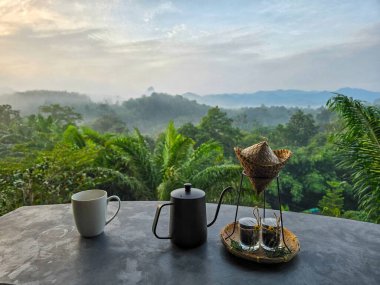 Khao Sok, Tayland 'da huzurlu bir sabahın tadını çıkarın. Yoğun yağmur ormanlarının üzerinde güneş doğuyor. Geleneksel bir çay seti, yemyeşil manzaralı ve sisli dağlarla dolu bir masada bekliyor..
