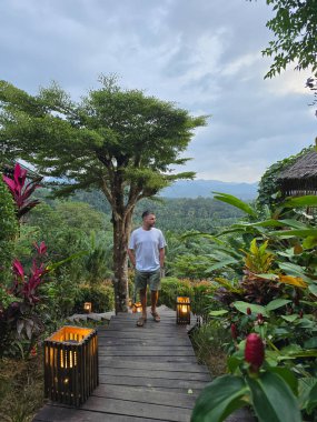 Tayland, Khao Sok 'ta canlı tropikal bitki örtüsüyle çevrili ahşap bir patika boyunca sakin bir yürüyüş. Alacakaranlık yemyeşil araziye çöktüğünde fenerler yolu aydınlatır.