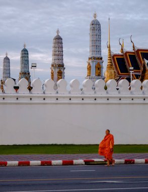 Bangkok, Tayland, 19 Kasım 2025. Gün batımının sıcacık parıltısıyla ışıldayan kuleleriyle görkemli Wat Phra Kaew 'da bir keşişin zarafetle yürüdüğü sakin bir manzara gözler önüne seriliyor..