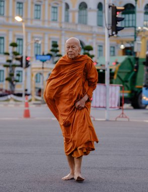 Bangkok, Tayland, 19 Kasım 2025. Geleneksel turuncu cüppeli sakin bir keşiş Wat Phra Kaew yakınlarındaki kalabalık bir caddede çıplak ayakla yürüyor..