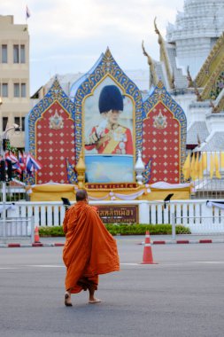 Bangkok, Tayland, 19 Kasım 2025, turuncu cübbeli sakin bir keşiş etkileyici Wat Phra Kaew 'in yanından geçiyor büyük bir kraliyet portresiyle süslenmiş. Atmosfer kültürel zenginliği yansıtıyor..