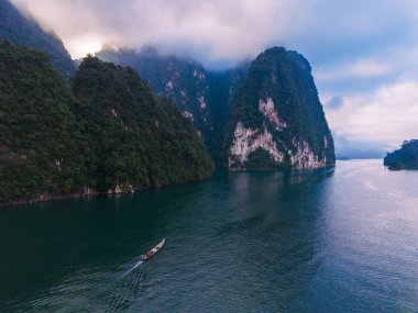 Khao Sok, Tayland 'daki Cheow Lan Gölü' nün sakin sularını deneyin. Zümrüt sularda süzülen küçük bir tekne, yemyeşil tepeler ve kasvetli bir gökyüzünün altındaki dramatik kireçtaşı kayalıklarıyla çevrili..