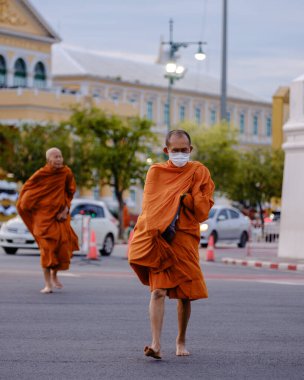 Bangkok, Tayland, 19 Kasım 2025. Canlı turuncu cüppeli keşişler Wat Phra Kaew yakınlarındaki sokaklarda dolaşıyorlar. Huzurlu atmosfer, tarihi mimarinin arka planıyla güçlenir..