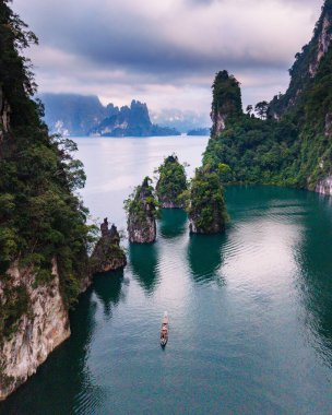 Tayland 'ın Khao Sok kentindeki Cheow Lan Gölü' nün sakin manzarasını deneyin. Görkemli kireçtaşı oluşumları ve yemyeşil yemyeşil alanlarla çevrili geleneksel tekneler zümrüt sularının üzerinde süzülüyor..