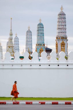 Bangkok, Tayland, 19 Kasım 2025, Wat Phra Kaew 'in beyaz duvarı boyunca yürüyen dingin bir keşiş, tapınağın canlı renklerini bulutlu bir gökyüzüne karşı sergiliyor..