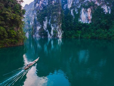 Uzun kuyruklu bir tekne, dramatik kireçtaşı uçurumları ve yemyeşil yeşilliklerle çevrili olan Cheow Lan Gölü 'nün sakin sularında süzülür. Khao Sok, Tayland