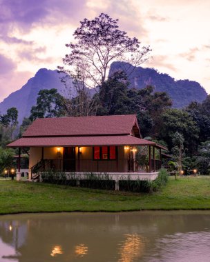 Büyüleyici bir kulübe, Tayland 'ın Khao Sok kentindeki yemyeşil yemyeşil ve dağlarla çevrili, deniz kenarında huzur içinde duruyor. Sihirli alacakaranlık gökyüzü ılık bir parıltı yayar, dingin bir atmosfer yaratır..