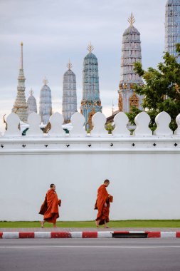Bangkok, Tayland, 19 Kasım 2025, turuncu cüppeli iki keşiş Wat Phra Kaew semtinde dolaşır, süslü kuleler ve yemyeşil alanlarla çevrili, bu tarihi alanın özünü yakalar..