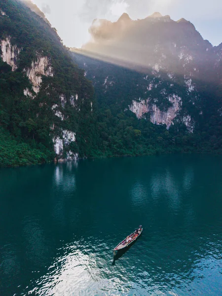 Khao Sok, Tayland 'daki Cheow Lan Gölü' nün dingin güzelliğini keşfedin. Küçük bir tekne, yüksek uçurumlar ve yumuşak güneş ışığı altında yemyeşil yemyeşil olarak çerçevelenmiş turkuaz suların üzerinde süzülür..