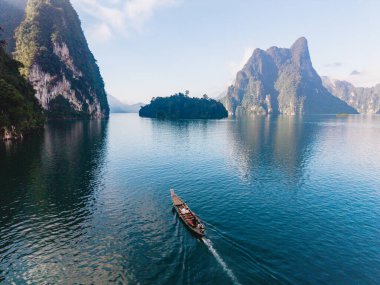 Geleneksel ahşap bir tekne, Khao Sok 'taki Khao Lan Gölü' nün kristal berrak sularında, görkemli kireçtaşı kayalıkları ve yemyeşil çimenliklerle çevrili bir şekilde süzülür..