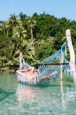Koh Kood Adası 'ndaki kristal berrak sularda asılı, verimli tropik bitki örtüsü ve palmiyelerle çevrili, Tayland' da güneşi ve sükuneti emen bir hamakta eğlenen bir adam.