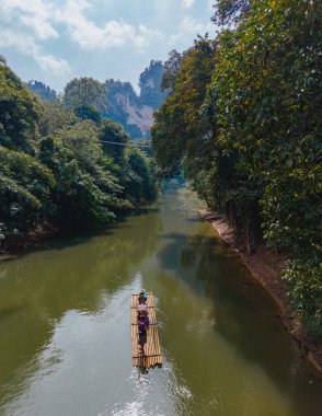 Tayland 'ın Khao Sok kentindeki iki gezgin, yoğun tropikal yapraklar ve yüksek kireçtaşı kayalıklarıyla çevrili bir bambu salında huzur içinde süzülüyor. Sakin su, parlak gökyüzünü yansıtır..