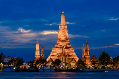 Wat Arun stands proudly against the evening sky, its intricate details glowing with warm lights. People explore the riverside, enjoying the beautiful atmosphere of Bangkok in twilight.