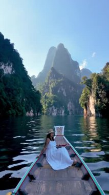Asyalı bir kadın, görkemli dağlar ve yemyeşil yeşilliklerle çevrili Khao Sok 'un sakin sularında, ahşap bir teknede dinleniyor. Huzurlu sahne Tayland 'ın doğal güzelliğini yansıtıyor..