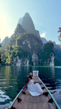 Tayland 'ın başkenti Khao Sok' un sakin güzelliğinin tadını çıkarın. Beyaz bir elbise içindeki bir kadın görkemli kireçtaşı kayalıkları ve kristal berrak sularla çevrili geleneksel bir teknede dinleniyor..