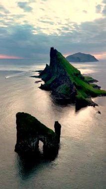 Dramatik kaya oluşumları, Faroe Adaları açıklarındaki sakin denizi delip geçer. Günbatımının sıcacık parıltısı manzaranın üzerine güzel renkler serpiştirir, çarpıcı bir görsel deneyim yaratır..