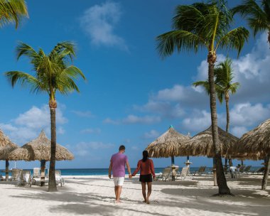 Çift, Palm Beach, Aruba 'nın yumuşak kumlarında, palmiye ağaçları ve büyüleyici sazanlı şemsiyelerle çevrili romantik bir yürüyüşün tadını çıkarıyor. Unutulmaz bir tatil için mükemmel bir tropikal kaçış..