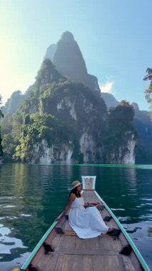 Bir gezgin, Khao Sok Ulusal Parkı 'nın çarpıcı zümrüt suları ve dramatik kireçtaşı uçurumlarıyla çevrili ahşap bir teknede dinlenir. Tayland doğasında sükunet deneyimi.