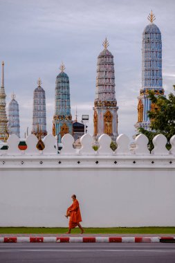 Bangkok, Tayland, 19 Kasım 2025, Wat Phra Kaew duvarı boyunca zarif bir şekilde yürüyen bir keşiş, gökyüzüne karşı ikonik süsleme kuleleri sergiliyor. Huzurlu sahne kültürel zenginliği yakalıyor..
