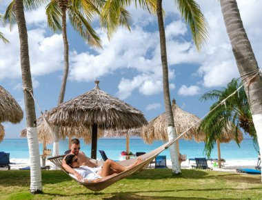 Bir çift, sallanan palmiye ağaçları ve plaj şemsiyeleriyle çevrili Divi Sahili Aruba 'daki bir hamakta huzurlu bir anın tadını çıkarıyor. Turkuaz sular ve yumuşak beyaz kumlar mükemmel bir tropikal kaçış yaratır..